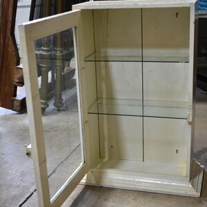 May include: A white wooden cabinet with a glass door and shelves. The door is open and the shelves are empty.