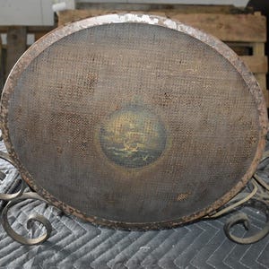 May include: A round, antique serving tray with a dark, textured surface and a decorative metal base. The tray features a small, circular painting of a ship on the surface. The metal base has ornate, scrolled details.