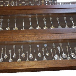May include: Three rectangular wooden display cases with glass lids, each containing a collection of silver-toned souvenir spoons arranged in a single row. The wooden frames have a medium brown finish, and the spoons feature various decorative handles.