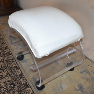 May include: A white upholstered ottoman featuring a smooth, padded top cushion. The base is constructed from clear, curved acrylic material, forming a C-shape on each side. The unit is supported by four small black caster wheels for mobility.