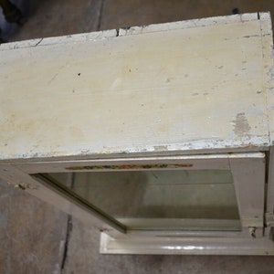 May include: A white wooden display case with a glass front and a decorative floral design on the top edge. The case has a simple, vintage style.