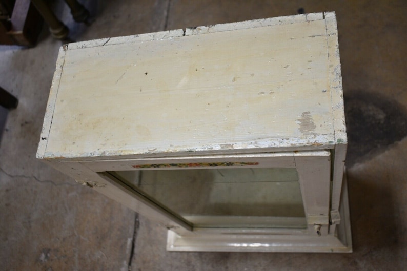 May include: A white wooden display case with a glass front and a decorative floral design on the top edge. The case has a simple, vintage style.