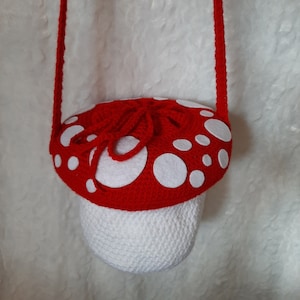 May include: A red and white crocheted mushroom-shaped bag with a white strap. The mushroom has white felt dots on the top.
