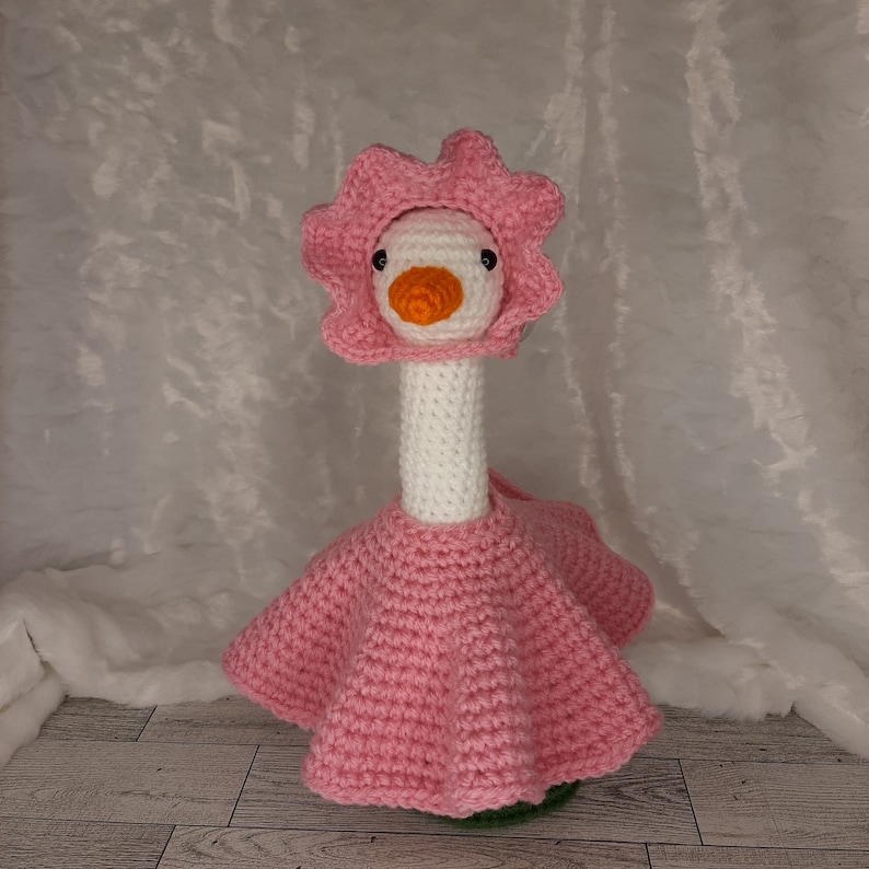 May include: A handmade crochet goose doll wearing a pink flower hat and dress. The goose has a white body, an orange beak, and black eyes. The pink dress has a flared skirt. The doll is standing on a green base.