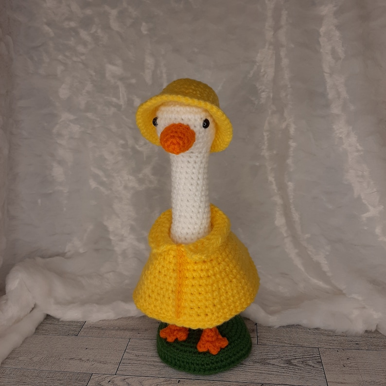 May include: A handmade crochet goose wearing a yellow raincoat and hat. The goose has a white body, orange beak and feet, and black eyes. It is standing on a green base.