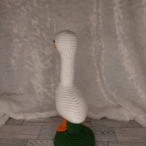 May include: A handmade crochet goose figurine. The goose is primarily white with an orange beak and feet, and black eyes. It stands on a green base. The craftwork is detailed, showcasing the texture of the yarn.