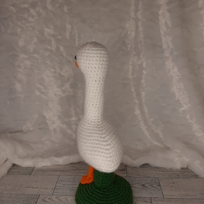 May include: A handmade crochet goose figurine. The goose is primarily white with an orange beak and feet, and black eyes. It stands on a green base. The craftwork is detailed, showcasing the texture of the yarn.