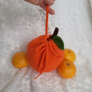 Pode incluir: Uma bolsa de cordão de crochê em forma de laranja com uma folha verde e um caule marrom. A bolsa está sentada em uma superfície branca e felpuda com três laranjas inteiras.