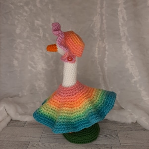 May include: A whimsical, handmade crochet goose figurine. The goose wears a rainbow-colored dress, a pink hat, and a matching collar. The goose has an orange beak and is standing on a green base. The overall design is colorful and cheerful.