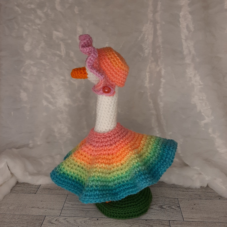 May include: A whimsical, handmade crochet goose figurine. The goose wears a rainbow-colored dress, a pink hat, and a matching collar. The goose has an orange beak and is standing on a green base. The overall design is colorful and cheerful.