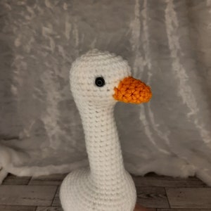 May include: A handmade crochet goose with a white body, long neck, and an orange beak. The goose has a black eye and is made of soft yarn. The craft project is a whimsical decorative item.
