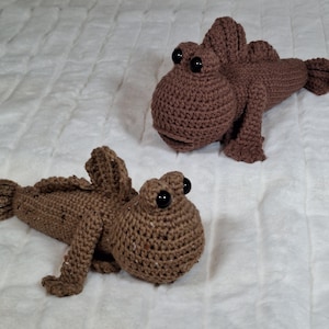 May include: Two brown crocheted mudskipper figures with large, round, black plastic eyes. Each creature features a textured, yarn-stitched body, prominent dorsal fins, and small pectoral fins. The figures are crafted with a consistent brown yarn.