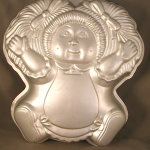 May include: A silver-colored metal cake pan shaped like a doll with pigtails and a bow. The doll is wearing a dress with a ruffle trim.