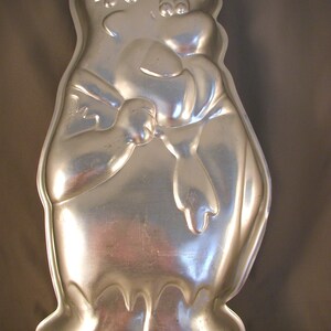 May include: A silver-colored, cartoon-style cake pan in the shape of a character with a large head, arms, and feet. The pan has a shiny, reflective surface and is designed to bake a cake in the character's likeness. The character has a smiling face and is holding its hands together.