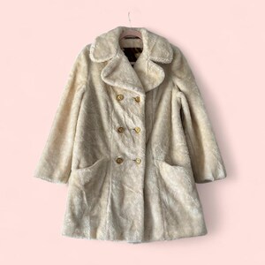 May include: Cream-colored faux fur coat with a double-breasted design. The coat features a wide collar, two front pockets, and gold-colored buttons. The coat is hanging on a pink hanger against a pink background.