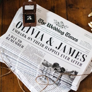 Pode incluir: Uma pilha de jornais de anúncio de casamento com a manchete "Olivia & James Embark on Their Happily Ever After" e uma foto do casal. O jornal está amarrado com barbante e uma pequena caixa de madeira com um anel dentro está no topo da pilha.