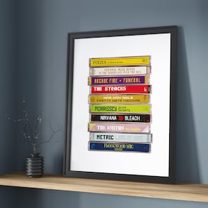 May include: A stack of vintage cassette tapes featuring various artists and album titles, including Pixies, Neutral Milk Hotel, Arcade Fire, The Strokes, Nine Inch Nails, Morrissey, Nirvana, The Smiths, Metric, and Fleetwood Mac. The tapes are arranged vertically in a white frame.