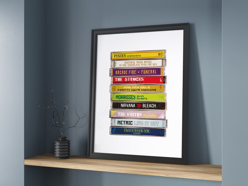 May include: A stack of vintage cassette tapes featuring various artists and album titles, including Pixies, Neutral Milk Hotel, Arcade Fire, The Strokes, Nine Inch Nails, Morrissey, Nirvana, The Smiths, Metric, and Fleetwood Mac. The tapes are arranged vertically in a white frame.
