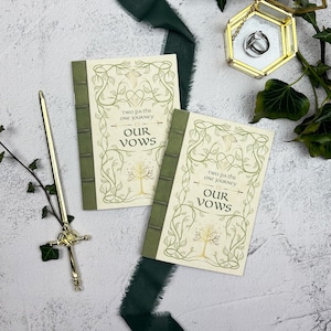 Può includere: Due libretti di promesse di matrimonio con dorso verde e copertine color crema. Le copertine presentano un motivo floreale decorativo e le parole "Two paths one journey" e "Our Vows". Una penna d'oro e un portagioie sono anche nell'immagine.