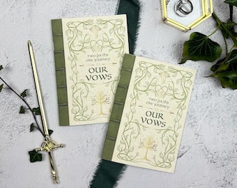 May include: Two wedding vow books with green spines and cream-colored covers. The covers feature a decorative floral design and the words "Two paths one journey" and "Our Vows". A gold pen and a ring box are also in the image.