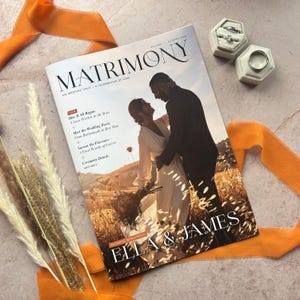 May include: A wedding magazine titled "MATRIMONY" with a cover photo of a couple embracing. The magazine features the text "ELLA & JAMES". Two ring boxes with rings are visible. Orange ribbon and dried plants are arranged around the magazine on a marble surface.