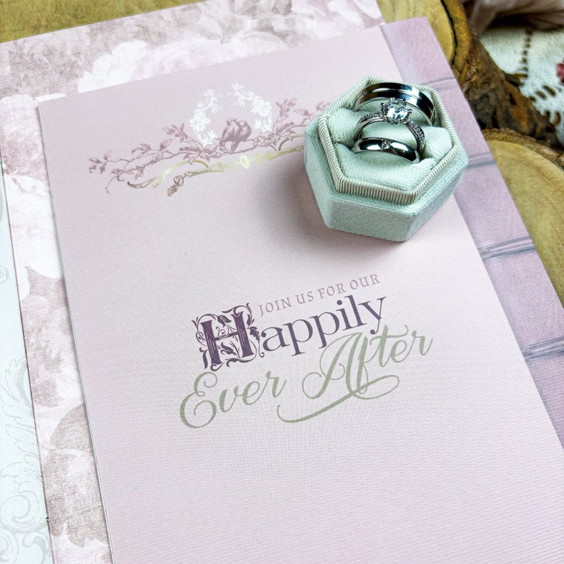 May include: A pink wedding invitation with the text "Join us for our Happily Ever After" in a decorative font. A small light blue velvet ring box with three silver wedding bands is on top of the invitation.