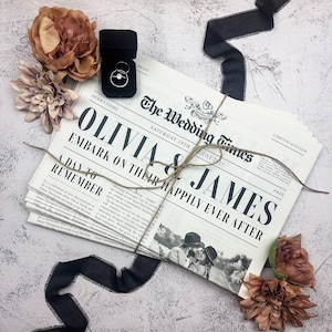 May include: A personalized newspaper titled The Wedding Times featuring the names Olivia and James, tied with twine. A black ring box containing two silver rings sits nearby, accompanied by three dusty rose-colored flowers and black silk ribbon.