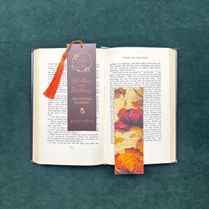 May include: Two rectangular bookmarks resting on an open book. One bookmark is dark brown with an orange tassel, featuring the text 'Save the Date', 'Willow and Anthony', 'ARE GETTING MARRIED', and '15.07.2023'. The other features a vibrant floral print.