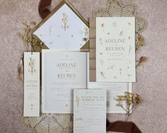 May include: A wedding stationery set featuring a book-style invitation, RSVP card, and bookmark, all printed with delicate wildflower illustrations. The text reads Adeline and Reuben, with dates and details in a neutral, cream-colored paper palette.