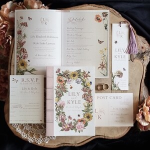 May include: Wedding invitation suite with floral designs. The invitation features a wreath of flowers with a sunflower, poppy, and other blooms. The RSVP card has a dragonfly and the text "R.S.V.P. Please return Lily & Kyle The West Cloister". The bookmark has the initials "L K" and the text "Lily & Kyle At The Cloister".