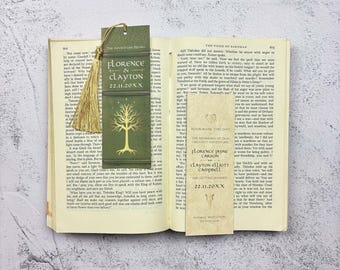 May include: Two rectangular paper bookmarks resting on an open book. One is dark green with a gold tree illustration and the text Florence & Clayton 22.11.20XX, featuring a gold tassel. The other is cream-colored with black text and a heart icon.