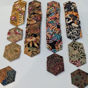 May include: A collection of fabric pieces in various floral and abstract patterns. The pieces are in a hexagonal shape, with colors including gold, brown, black, orange, and green. The fabric appears to be cut for quilting or other craft projects.