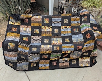 May include: A handmade quilt with a patchwork design. The quilt features squares of black fabric framing various animal print patterns in shades of brown, yellow, and blue. The quilt is draped over a surface, showcasing its size and design.