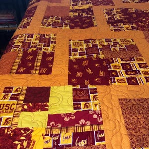 May include: A patchwork quilt featuring a repeating pattern of USC Trojans collegiate fabric in shades of cardinal red and gold, interspersed with solid gold fabric panels. The entire quilt is finished with a continuous heart-shaped quilting stitch.