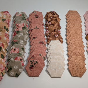 May include: Arrangement of fabric hexagon pieces in various floral and patterned designs. Colors include tan, pink, brown, and gray. The pieces are neatly stacked, ready for a quilting or crafting project.