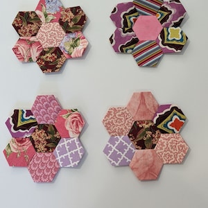 May include: Four hexagonal fabric flower blocks in shades of pink, purple, brown, and white. Each block is made of smaller hexagonal pieces of fabric sewn together.