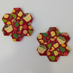 May include: Two red hexagon-shaped fabric coasters with a pattern of fruit, birds, and baskets. The coasters are made of fabric and have a quilted appearance.