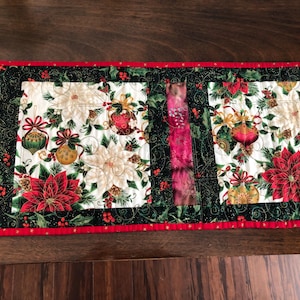 May include: A quilted table runner with a Christmas theme. It features a white background with red poinsettias, green leaves, and gold ornaments. The runner has a black and green border with a red trim.