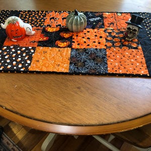 May include: A rectangular quilted table runner featuring a patchwork design of orange and black fabrics with Halloween motifs including pumpkins, spiderwebs, and candy corn. A ceramic ghost pumpkin, a small green pumpkin, and a skull are placed on top.