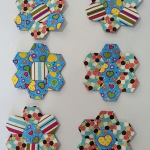 May include: Six colorful fabric hexagons with a flower-like design. Each hexagon is made of smaller hexagons in various patterns, including polka dots, stripes, and hearts. The hexagons are arranged in two rows of three.