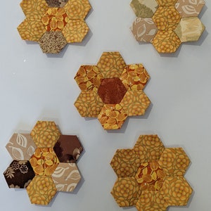 May include: Five hexagonal fabric flower-shaped coasters in shades of brown, yellow, and orange. The coasters are made of patchwork fabric with different floral patterns.