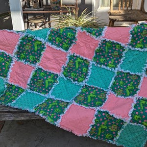 May include: A handmade patchwork quilt featuring a repeating pattern of green, pink, and teal squares. The green squares have a dinosaur print. The quilt has a white frayed edge and is displayed on a wooden bench.