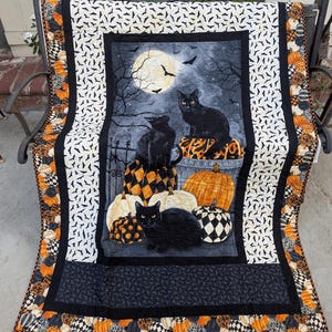 May include: A rectangular quilt featuring a central panel with three black cats sitting among orange, white, and black patterned pumpkins under a full moon. The border displays a repeating bat pattern and a decorative edge of colorful pumpkins.