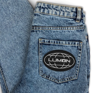 May include: Light blue denim jeans with a black and white patch on the back pocket. The patch features the word "LUMON" in white, with a globe design. The jeans have a classic five-pocket design.