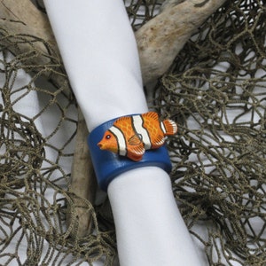 May include: A blue leather napkin ring with a hand-painted orange and white clownfish. The fish is detailed with black accents. The napkin ring is wrapped around a white cloth napkin, with a fishing net in the background.