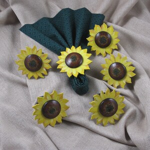 May include: Six sunflower napkin rings in yellow and brown with green fabric napkins. The napkin rings are made of wood and painted to look like sunflowers.