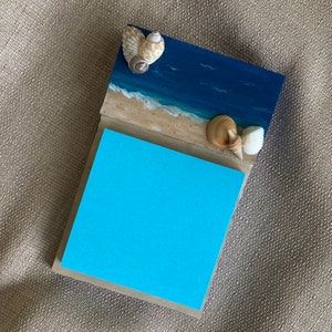 May include: A blue and beige wooden notepad with a seashell design. The notepad has a blue sticky note attached to it.