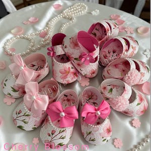 May include: A collection of white cardstock shoes decorated with pink cherry blossoms and bows. The shoes have pink interiors and are arranged on a white surface with scattered petals and a pearl necklace. Text on the image reads "Cherry Blossom Theme Cardstock Shoes."