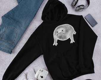 Puede incluir: Sudadera con capucha negra con un gráfico blanco de una rana frunciendo el ceño. La rana está dibujada en un estilo de dibujos animados con un cuerpo redondo y ojos grandes.