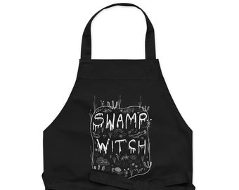 Op de afbeelding: Zwarte keukenschort met een wit ontwerp dat "Swamp Witch" zegt, omringd door moeras-thema illustraties.